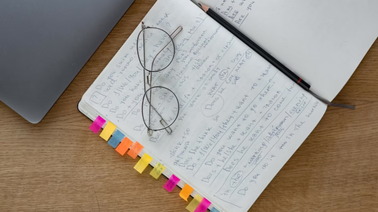 Top view of a notebook with handwritten notes, eyeglasses, and a pencil on a wooden desk.